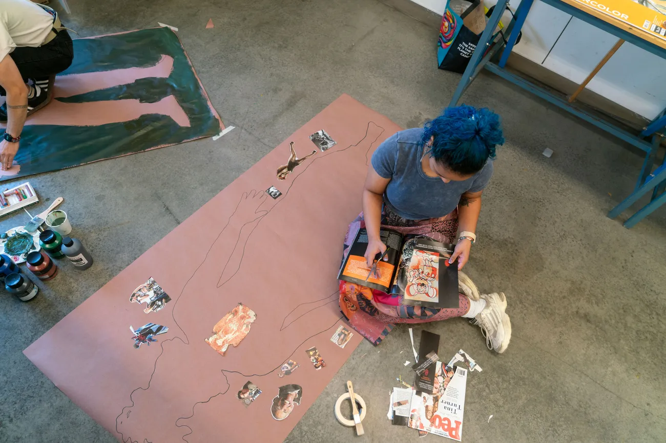 A student sits on the floor cutting pictures out of a magazine next to a full-size outline of a body.