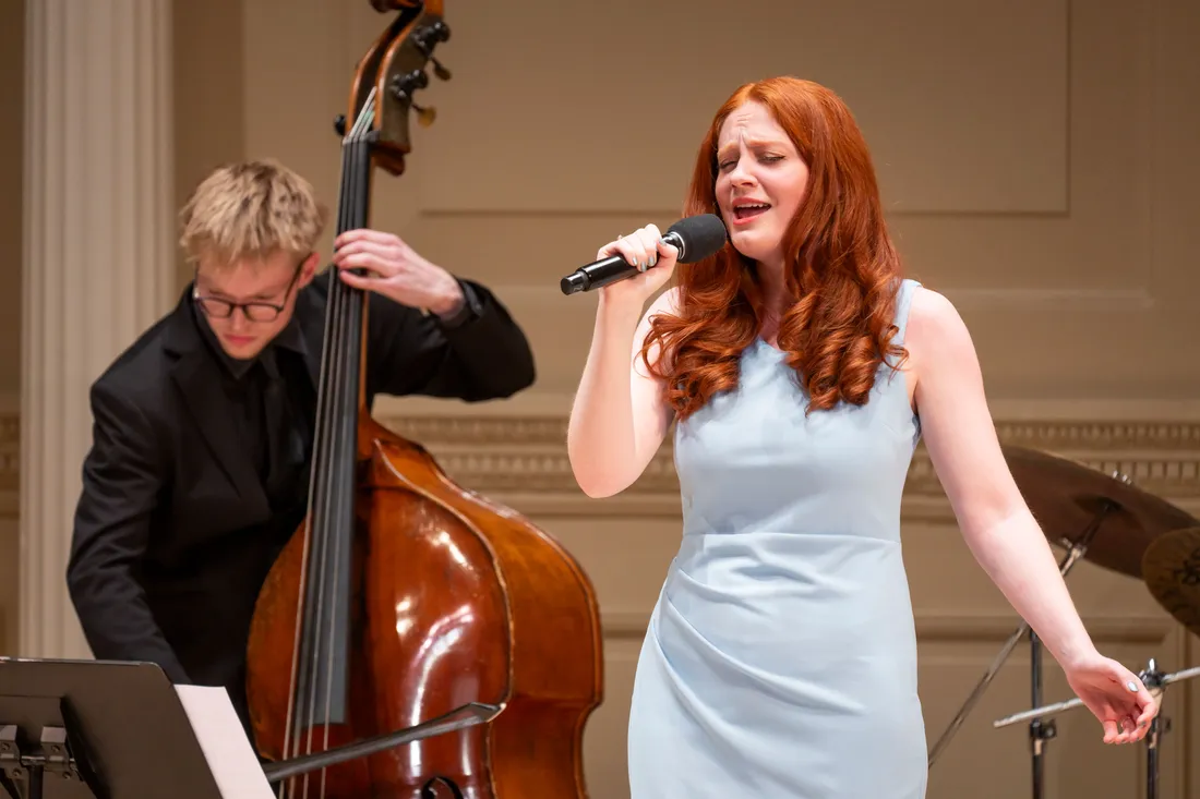 A person dressed in formal attire sings while another person plays bass in the background.