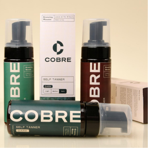 A collection of COBRE self-tanning mousse products arranged on a light beige background, featuring green and brown pump bottles, a white product box, and a horizontal bottle in the foreground.