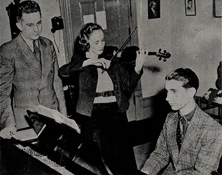 A student plays a violin while another plays a piano. A teacher looks on.