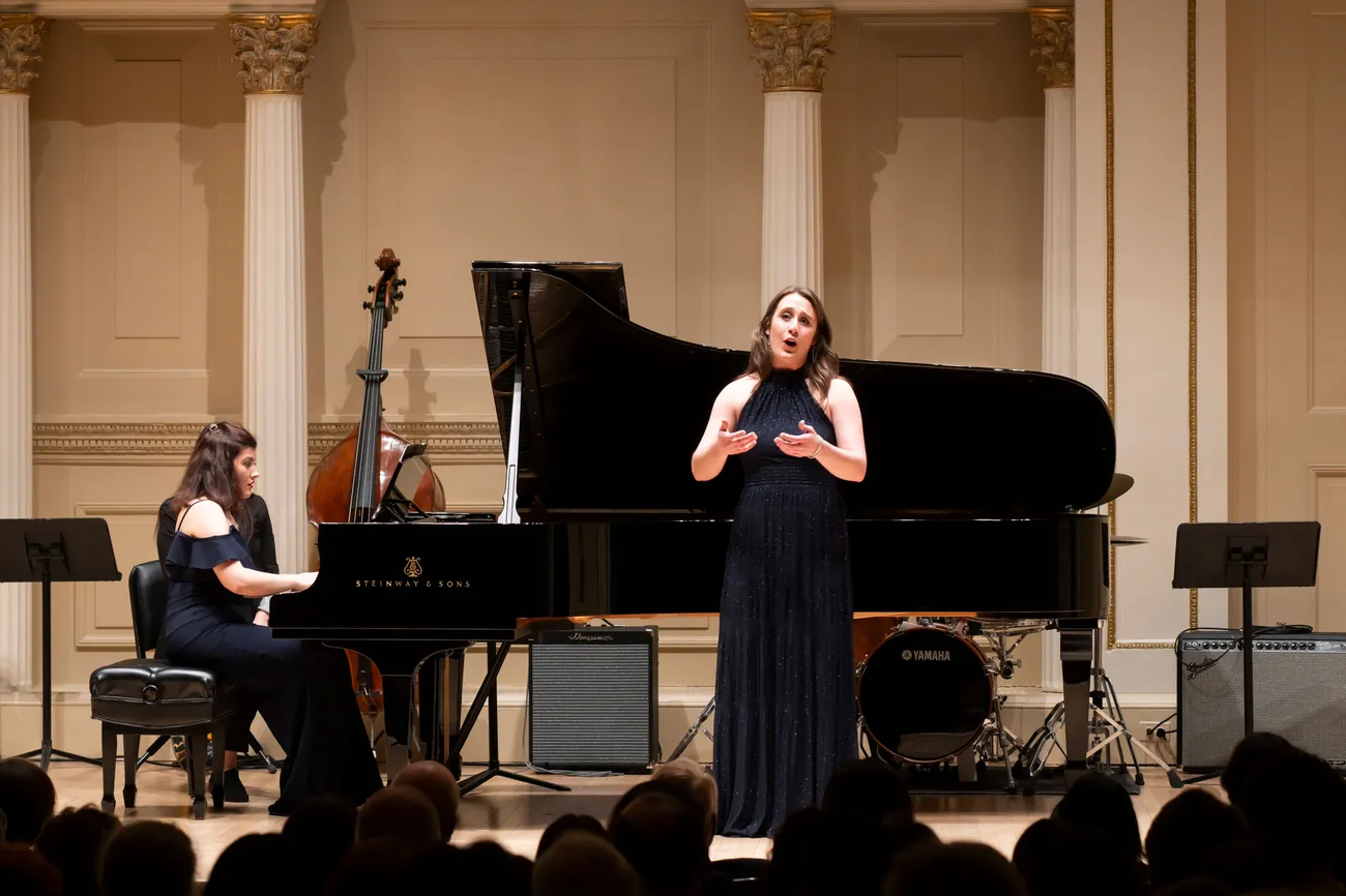A person stands on a stage in front of a piano singing. A person is playing the piano. Both are formally dressed.