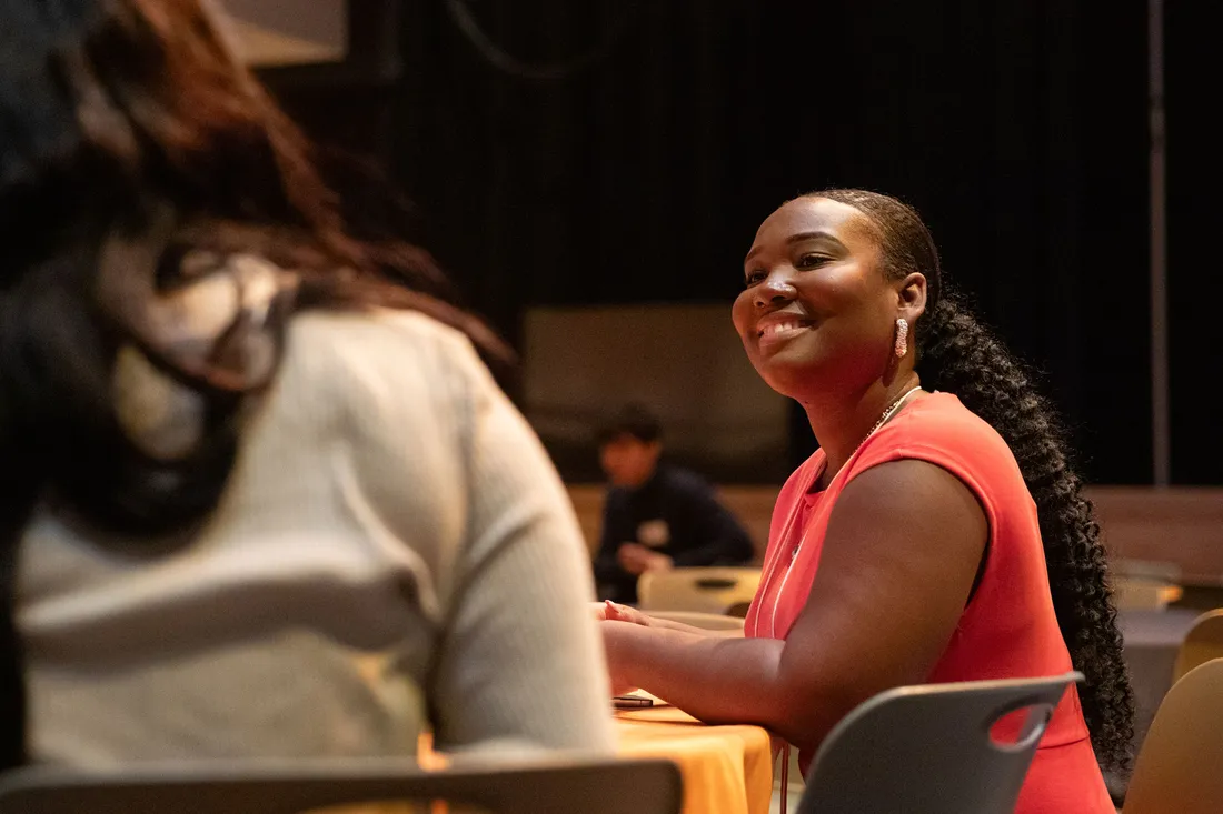 Two people sit at a table. One person's back is to the camera. The other person is smiling.