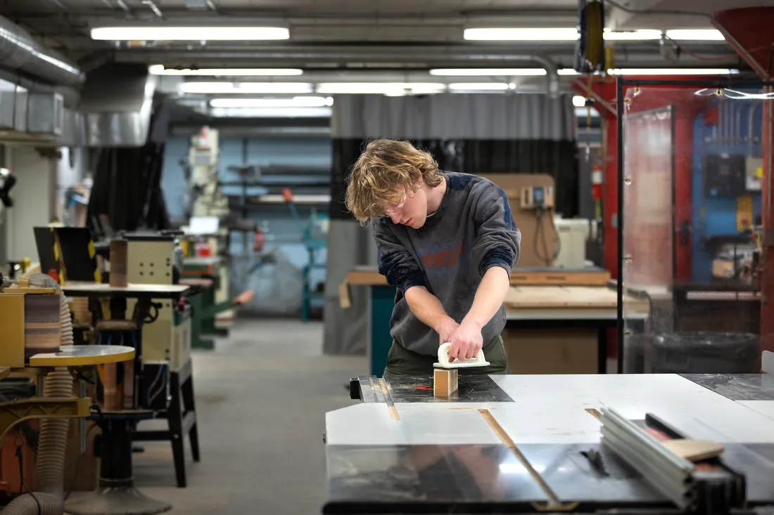 A student uses a woodworking tool on a table in a wood shop.