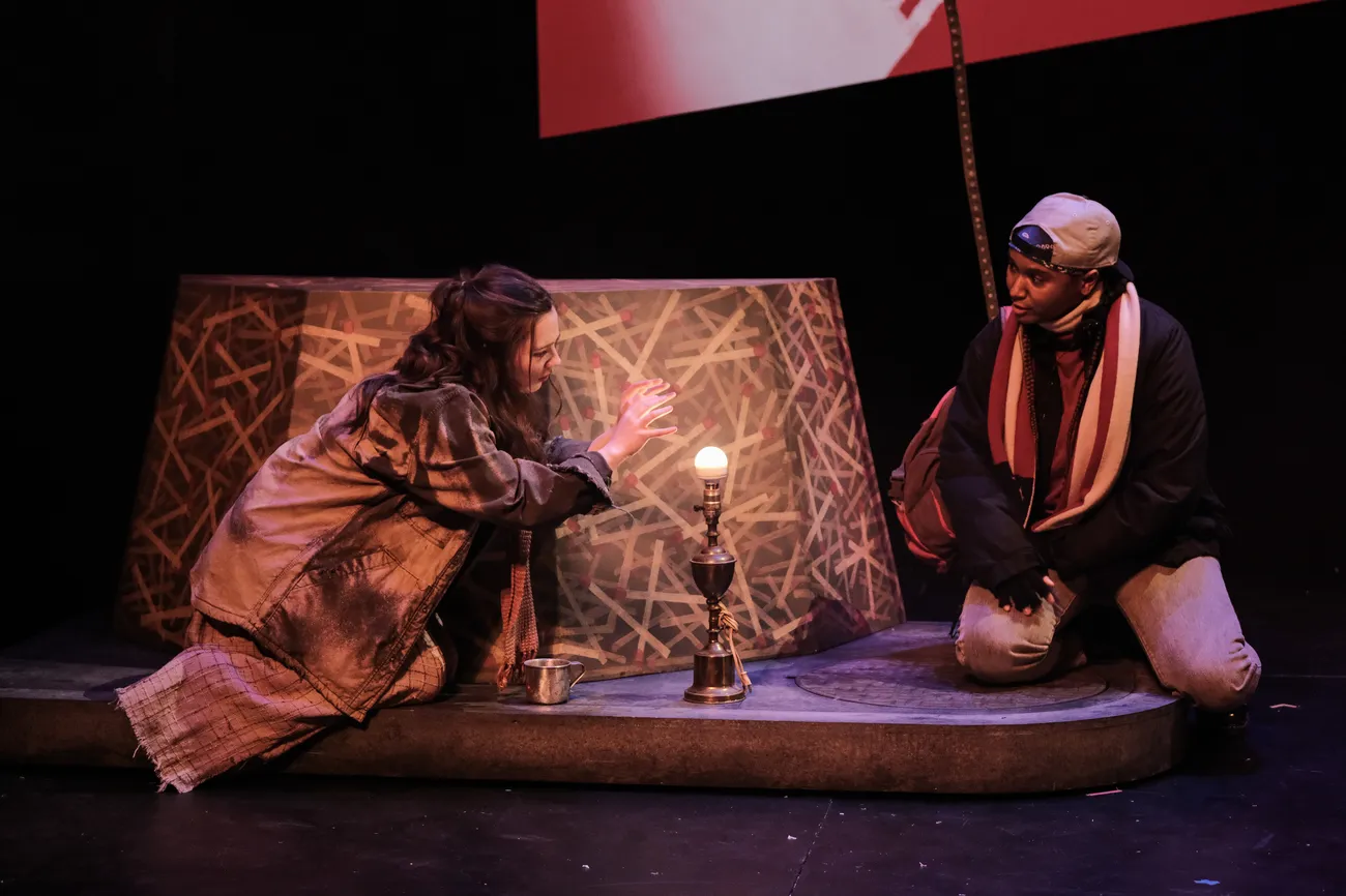 Two actors in costume sit on a stage with a lamp between them. The lamp does not have a shade.