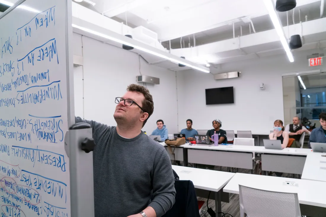 A person stands at a whiteboard and writes on it in front of a group of students who are sitting at a table.