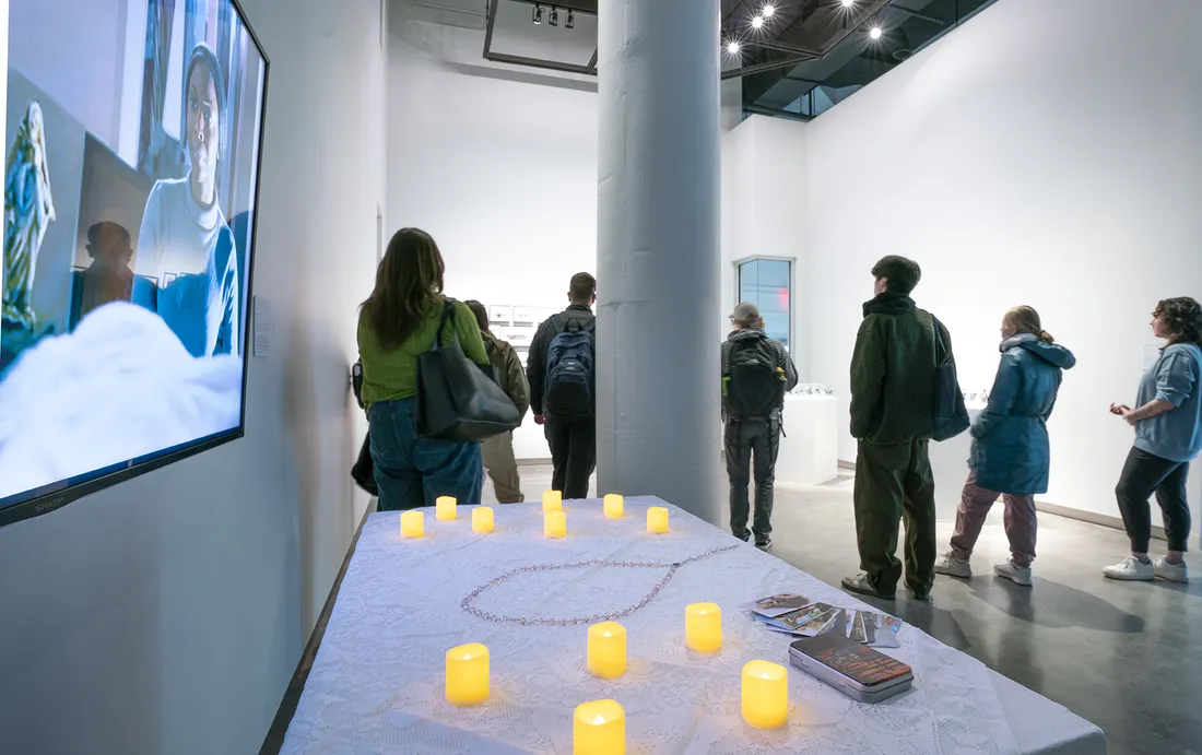 People stand and look at artwork in a gallery. On a wall is a screen playing a video of work, with lights on a table in front of it.