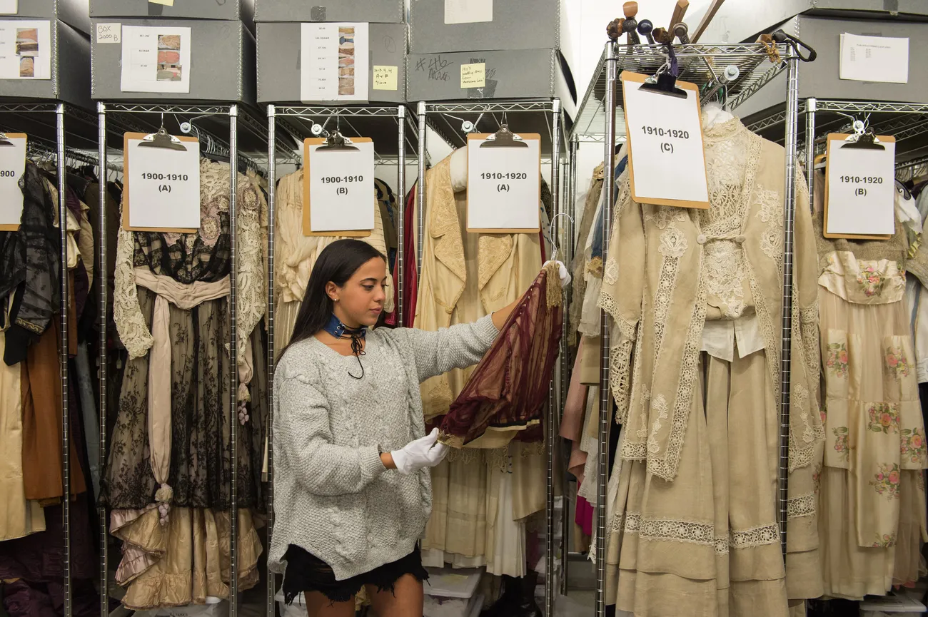 A student handles a garment wearing white gloves. Behind the student hangs racks and racks of dresses labeled by year.