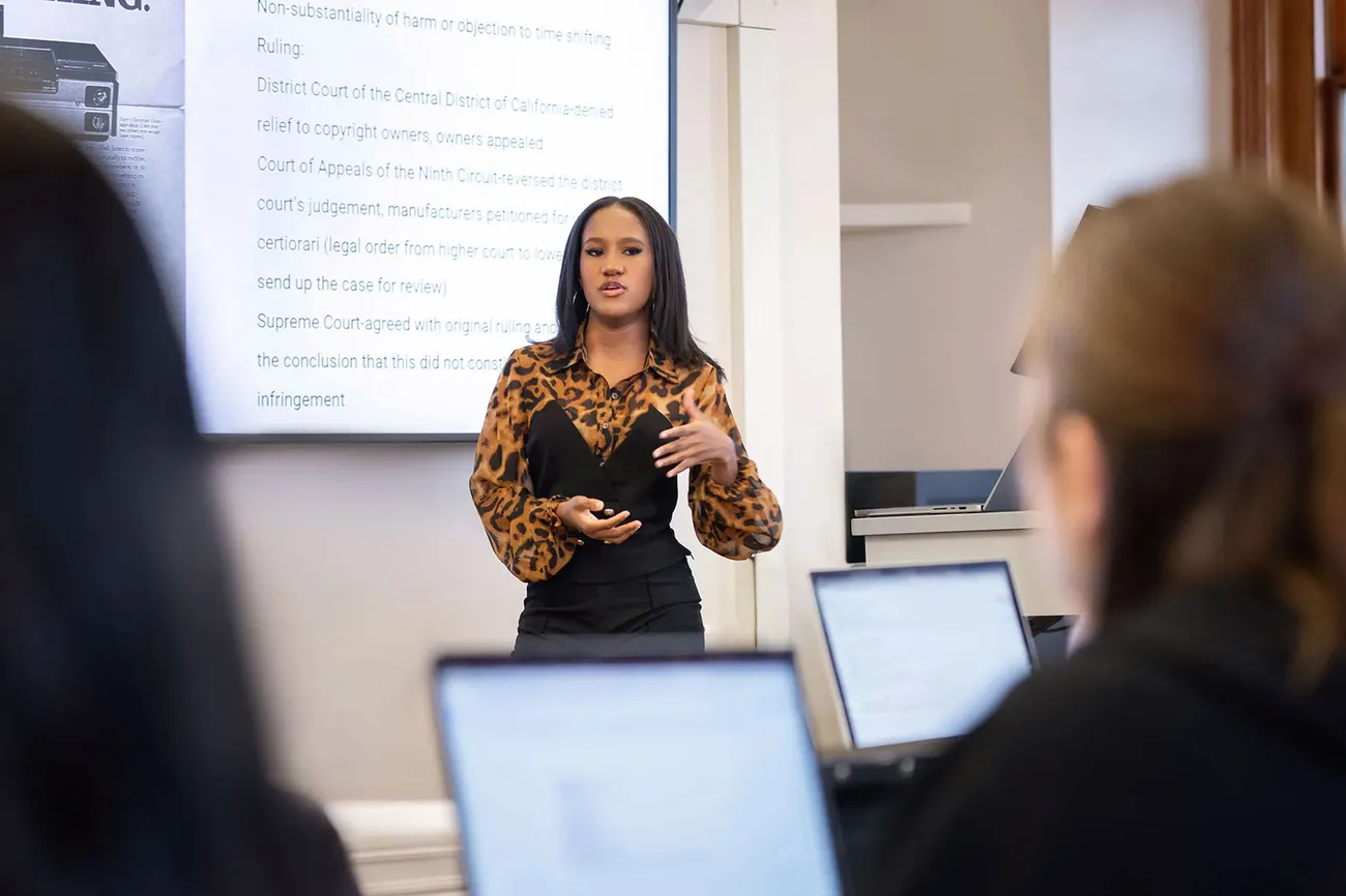 A student stands in front of a class talking and standing next to a screen.