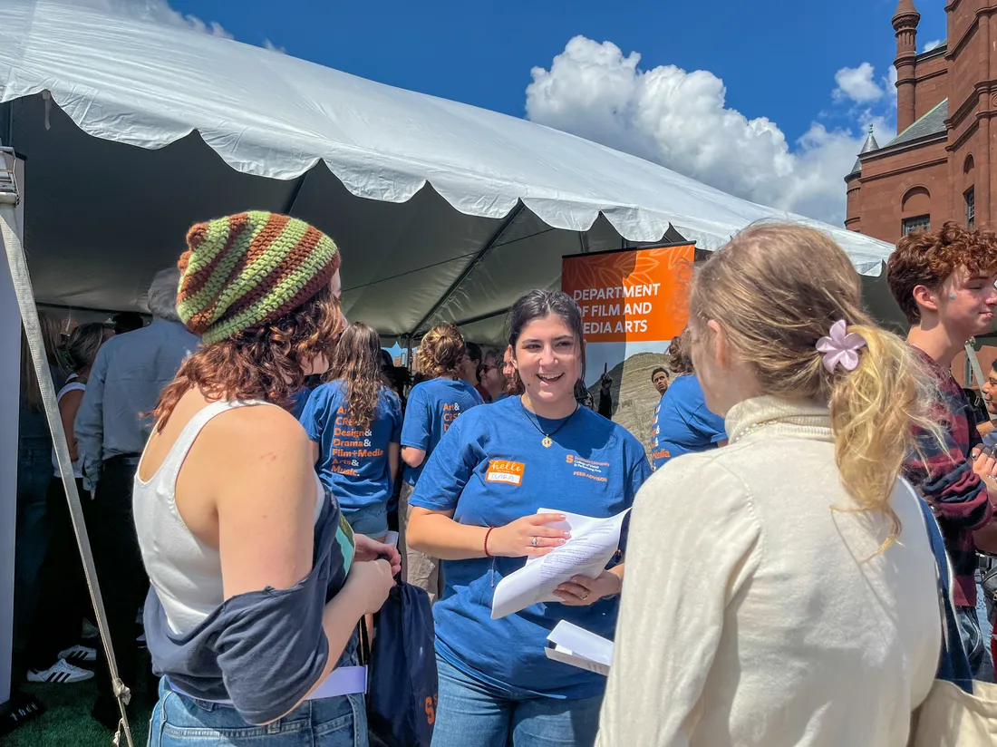 Three students talk outside a tent on a sunny day.