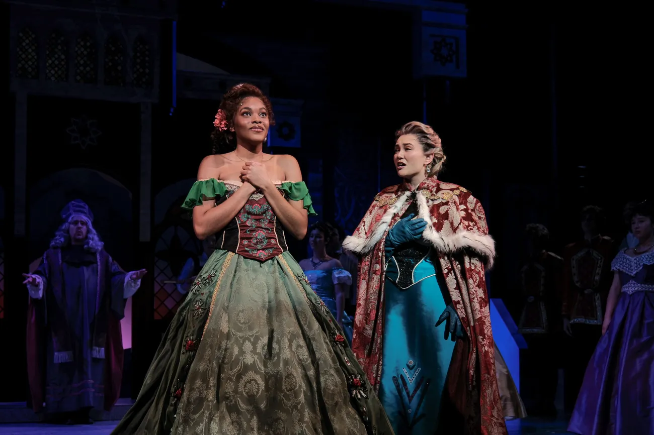 Two actors in ball gowns act a scene from Disney's "Frozen" on a dimly lit stage.