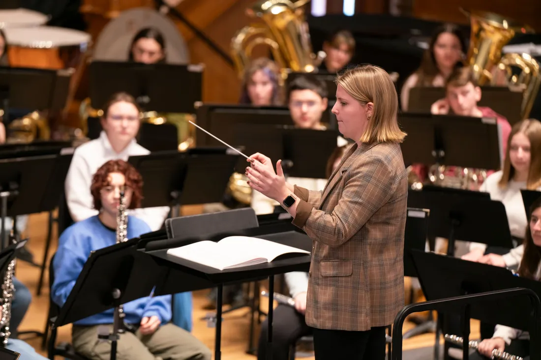 A student conductor stands in front of a wind ensemble with baton raised.