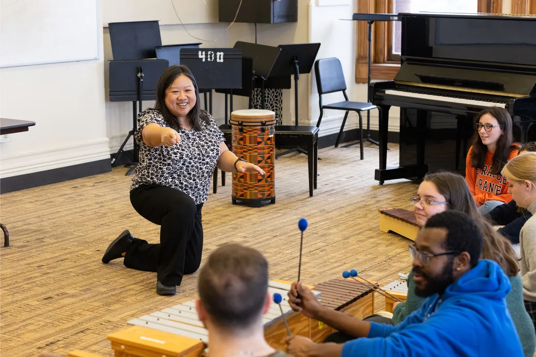 A faculty member kneels in front of a group of students sitting on the floor and gestures to them. They are holding mallets.