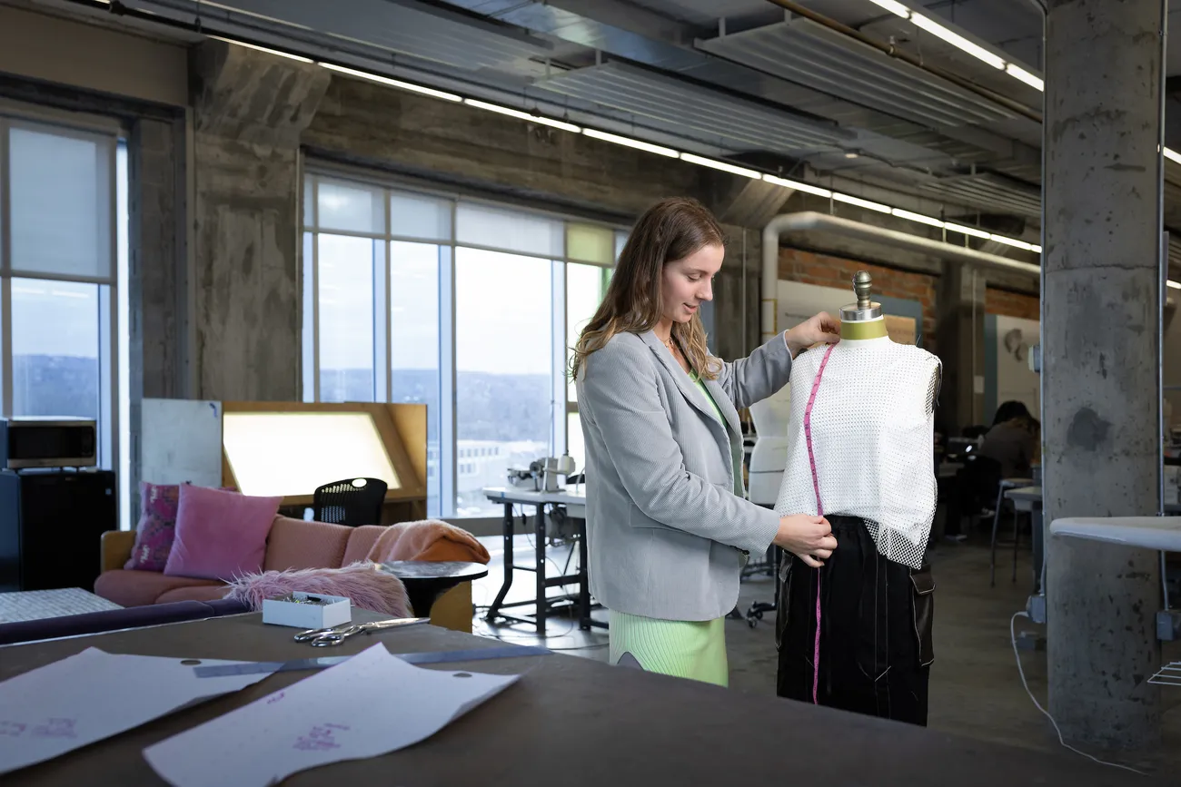 A student stands next to a mannequin measuring the garment hanging on the mannequin.