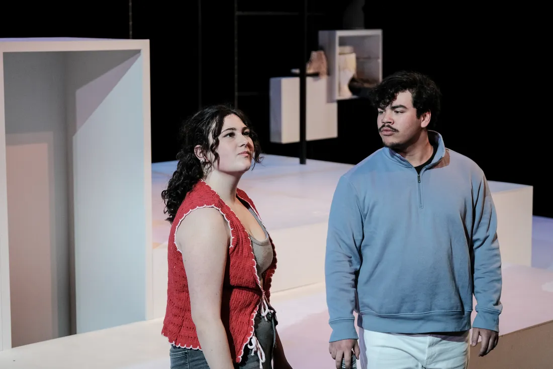Two students in costume act out a scene on a stage sparsely decorated in a set painted white.