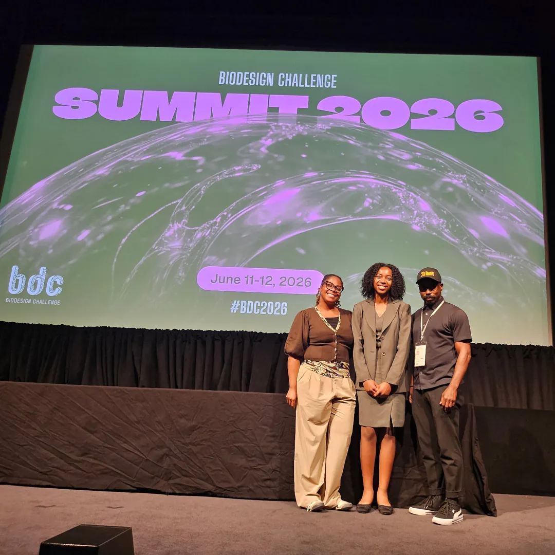 Three people stand in front of a screen that reads “Biodesign Challenge Summit 2026.”