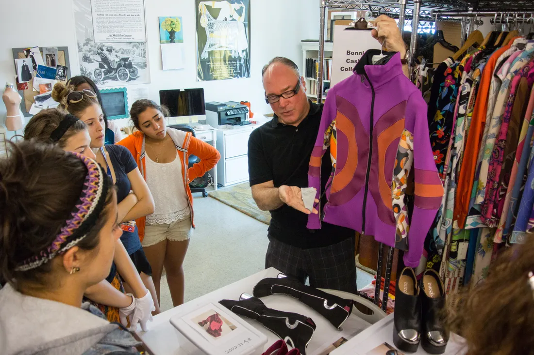 A faculty member holds up a pink jacket and talks to a group of students. They are standing in a room with many garments hung on racks.