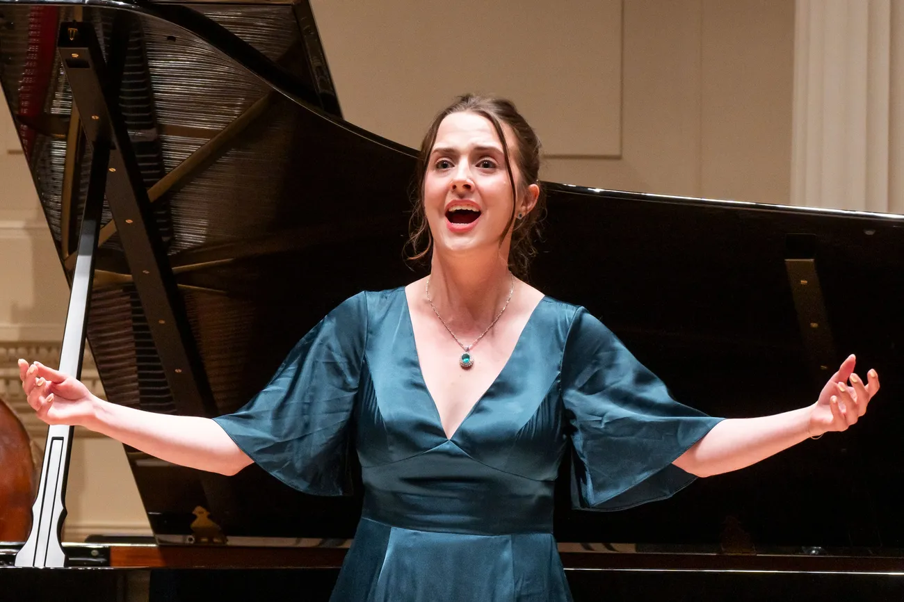 A person formally dressed sings on a stage in front of a piano.