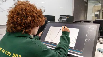 A student sits at a computer in a computer lab and uses a stylo to draw circular lines on the screen.