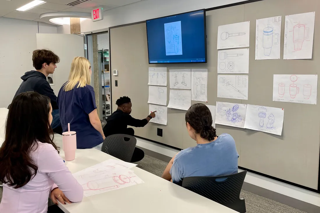 A faculty member points to a wall with designs pinned to it as a group of students listens.