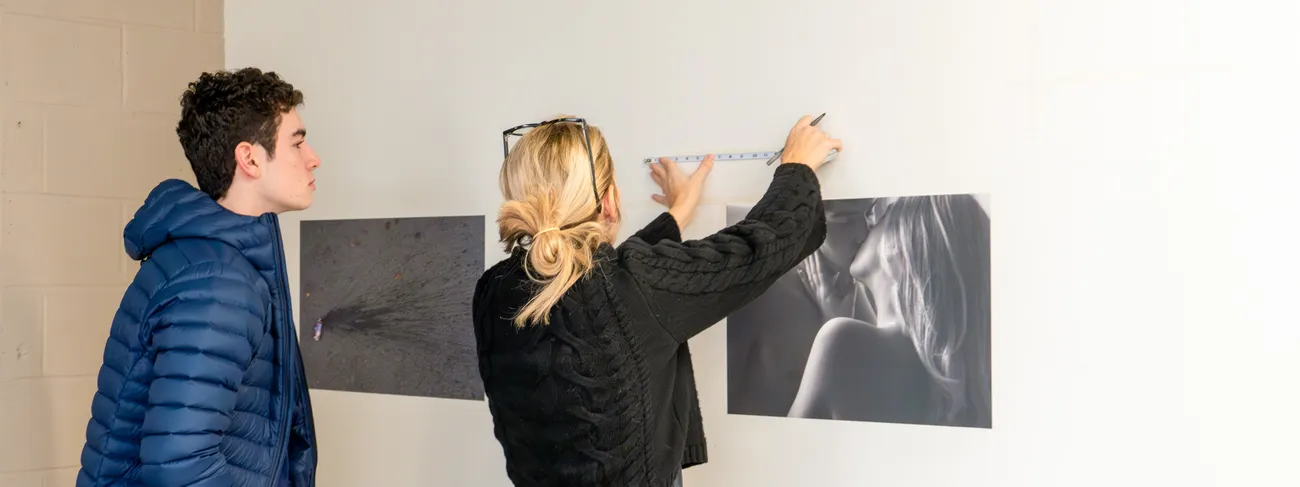 A student and faculty member hangs art on the wall.