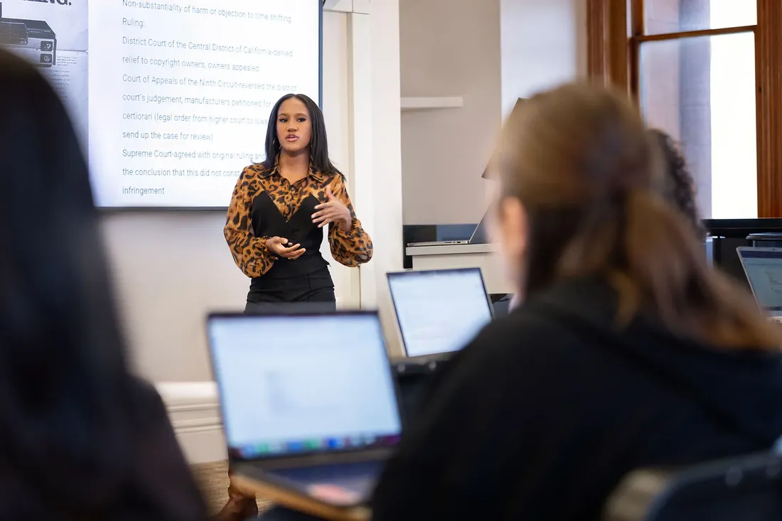 A student stands and gestures at the head of a class. Next to the student is a large projection screen with text about a Supreme Court ruling.