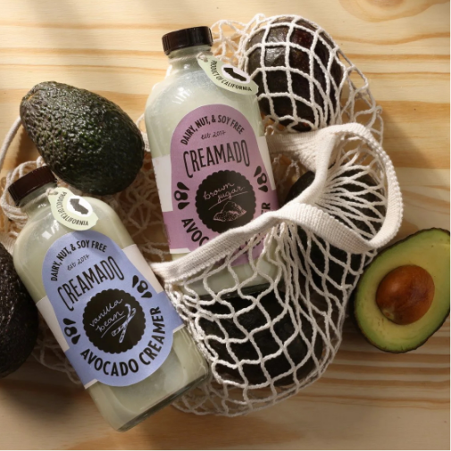 Two bottles of “Creamado” avocado creamer displayed inside a white mesh produce bag alongside whole and halved avocados on a light wooden surface. The packaging highlights dairy-, nut-, and soy-free ingredients with soft pastel labels and a natural, organic aesthetic.