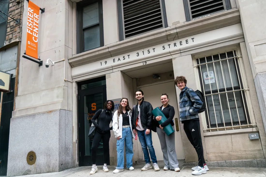 Five students pose and smile outside a building in New York City that has an orange sign that says Fisher Center.