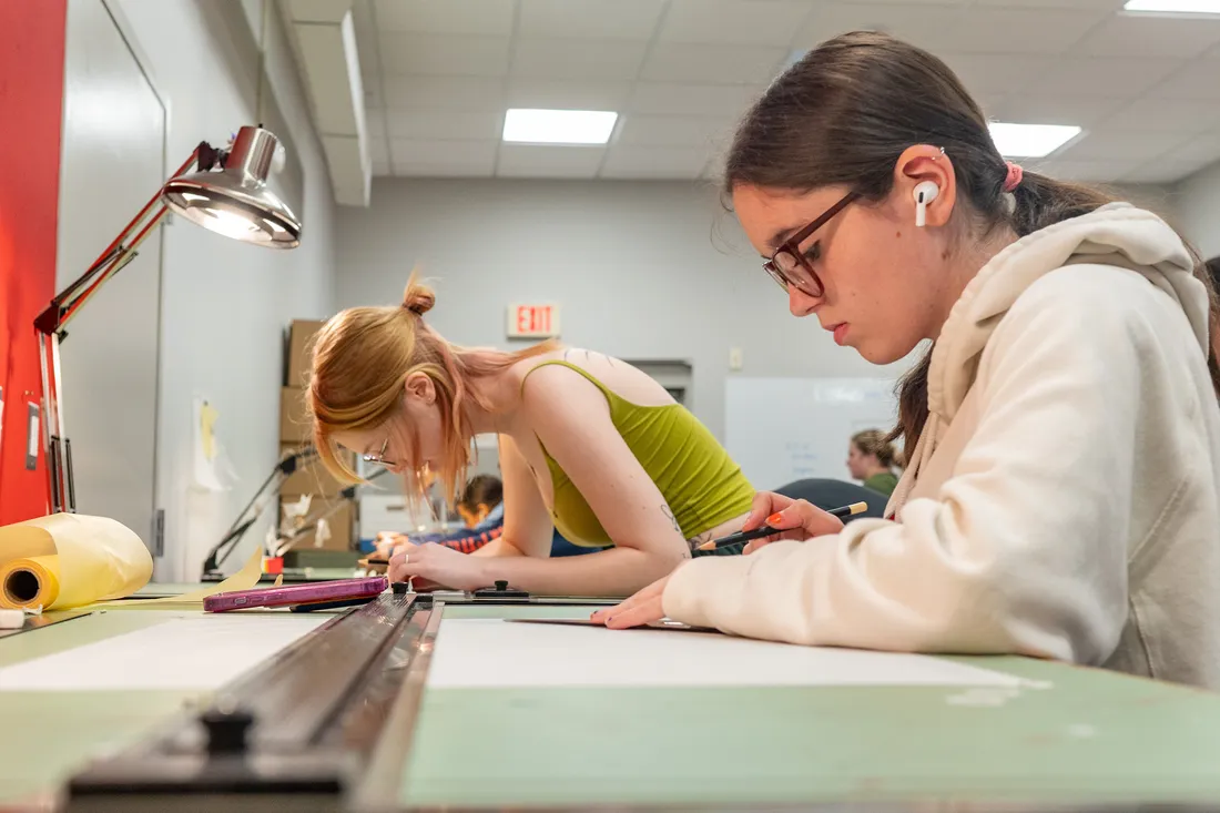 Two students lean over a drafting table and draw with a pencil and ruler.