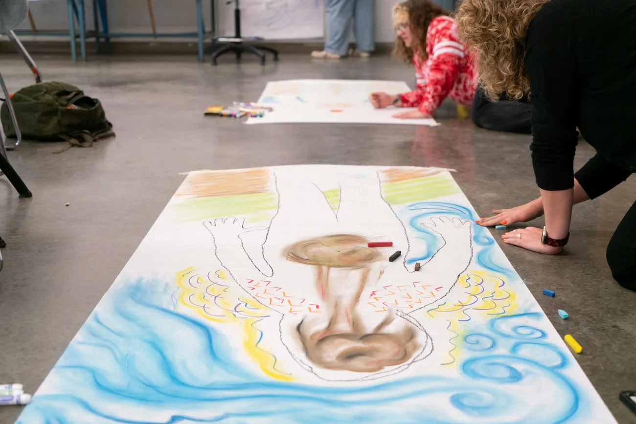 A student sits on the floor drawing on a large sheet of paper.