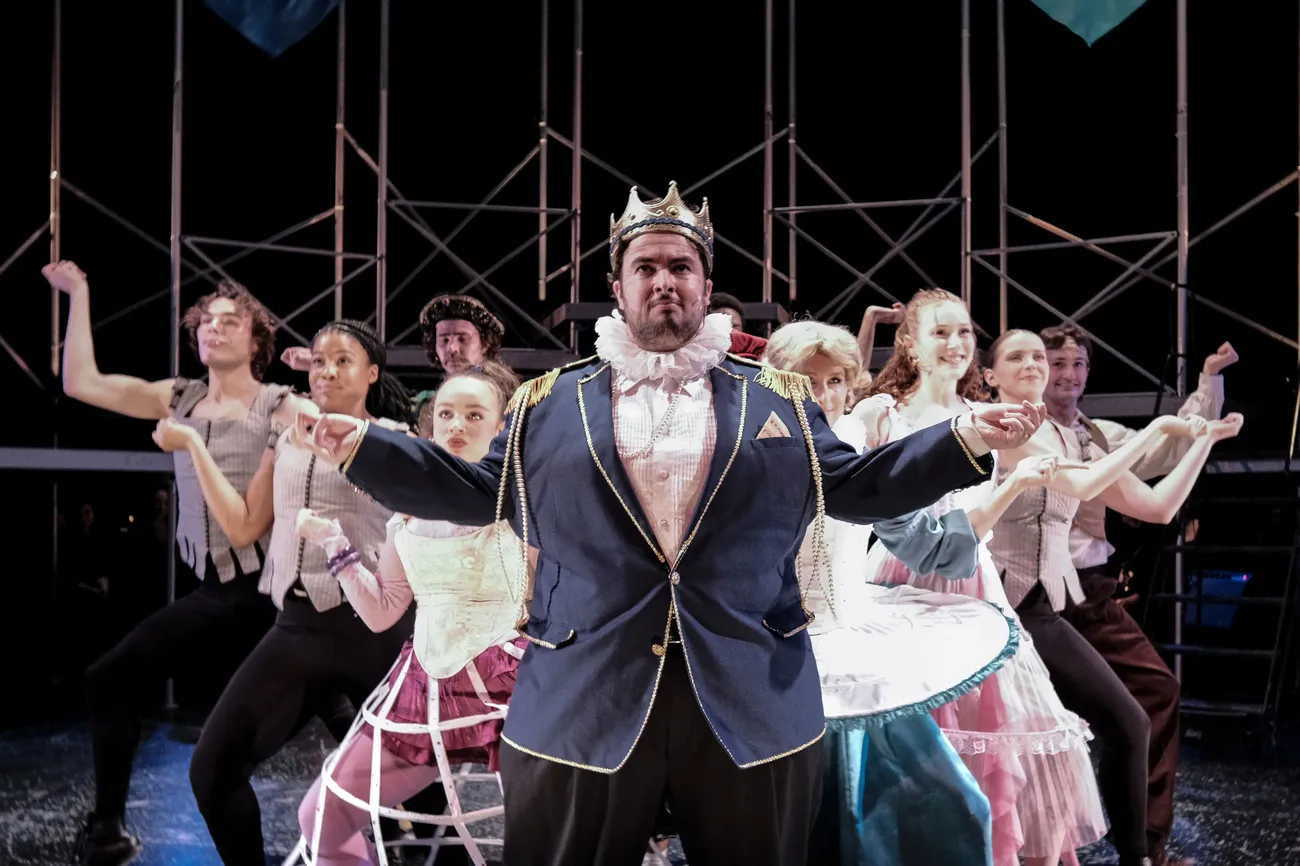 A group of actors in costume perform a dance number on a stage.