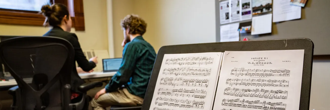 In the foreground is a music stand with a piece of music on it. In the background is two people sitting at a desk.