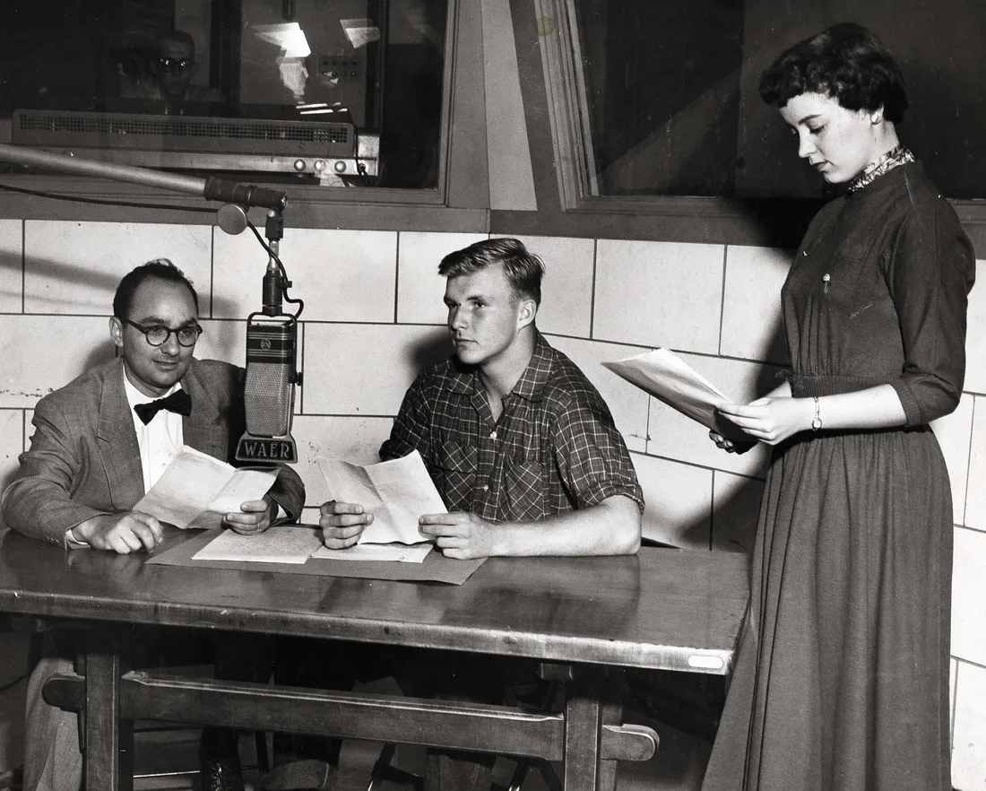 Three people sit and stand around the microphone in a radio studio in the 1940s.