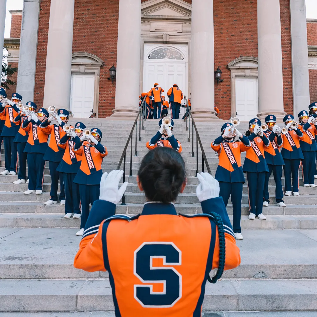 A person conducts the marching band on the steps of Hendricks Chapel.