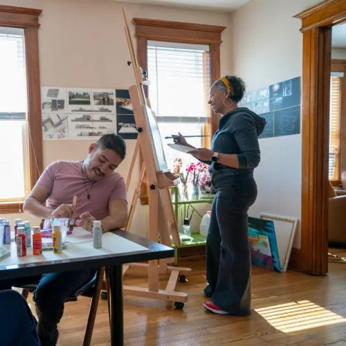A student works at a table. Professor Rochele Royster paints on a canvas in the background.