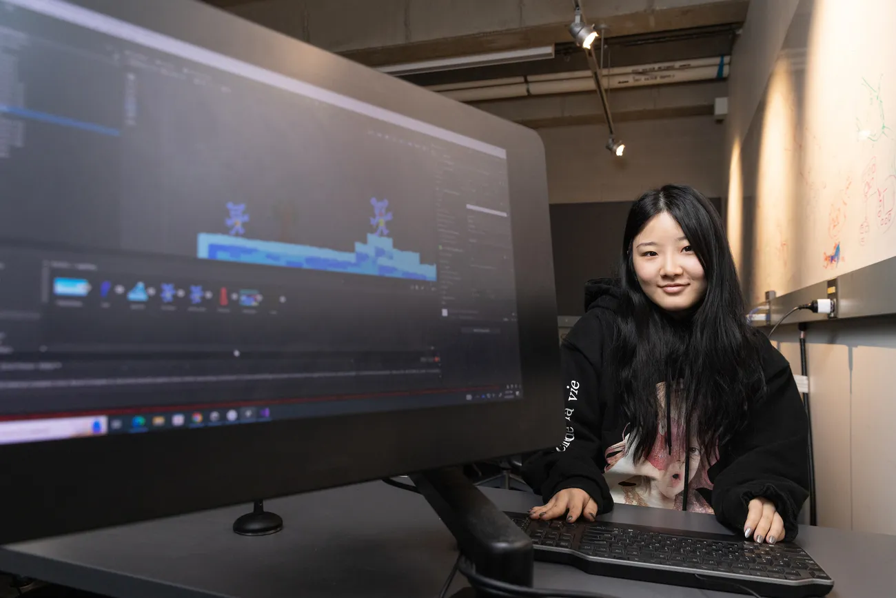 A student sits next to a computer screen that displays a computer game.