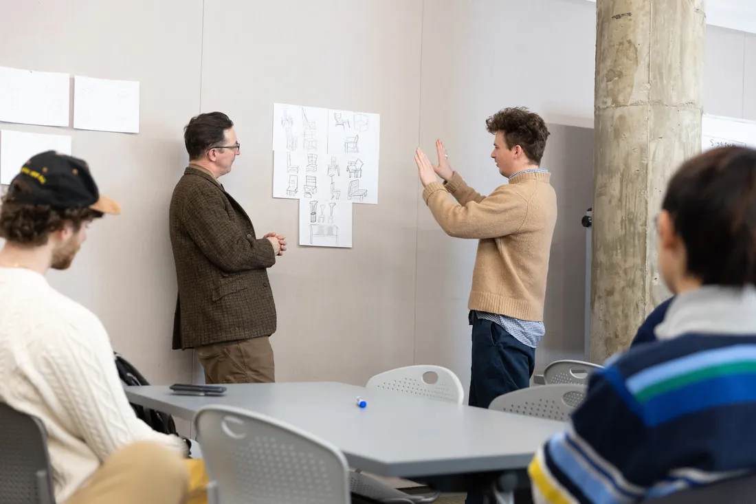 A teacher and a student stand at a wall looking at designs pinned on the wall while other students sit at desks.