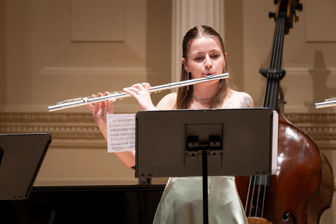 A student formally dressed stands behind a music stand playing the flute.