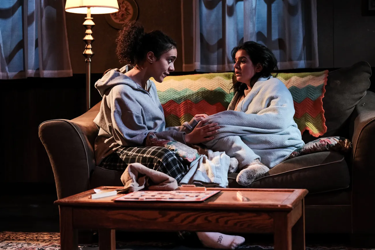 Two actors in contemporary costumes sit on a couch and talk.