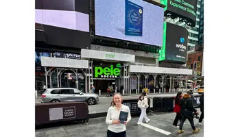 Laurie Maddalena stands on a street holding her book; behind her is a screen showing her book.