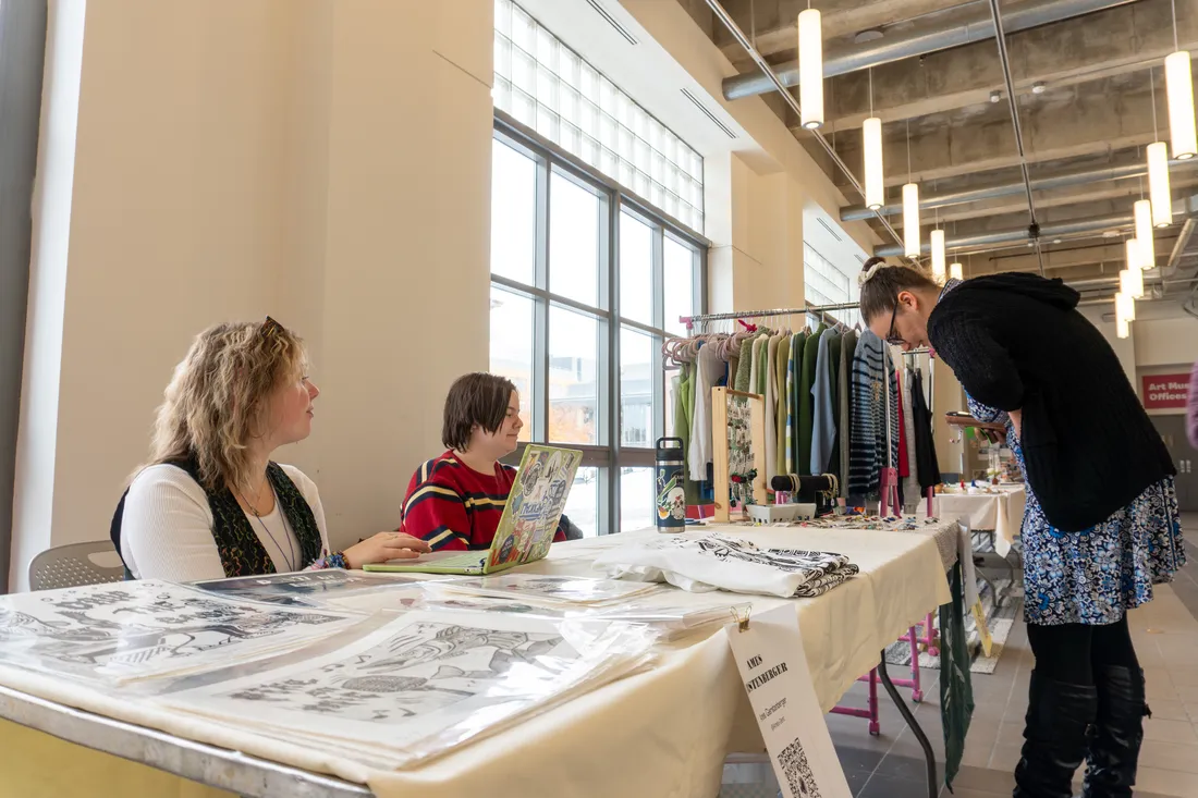 Two students sit at a table that has prints displayed while a person stands and looks at them.
