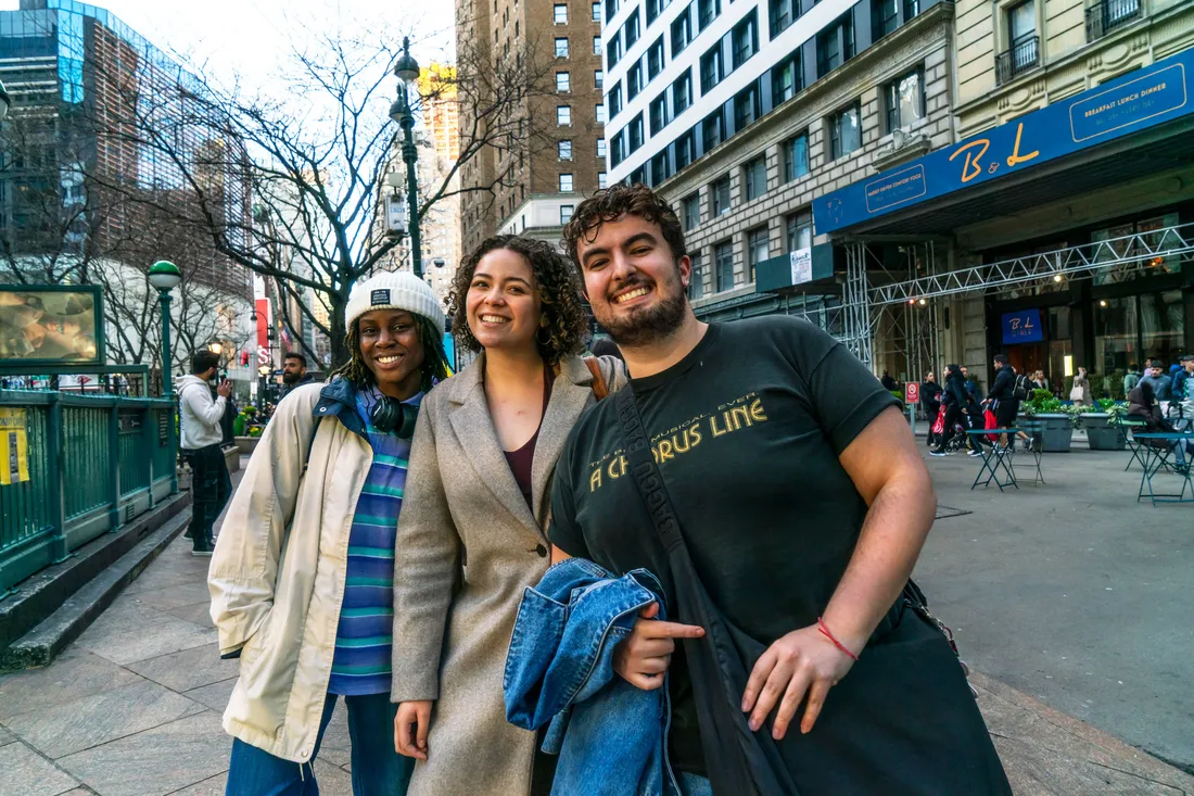 Three students pose and smile on a busy street in New York City.