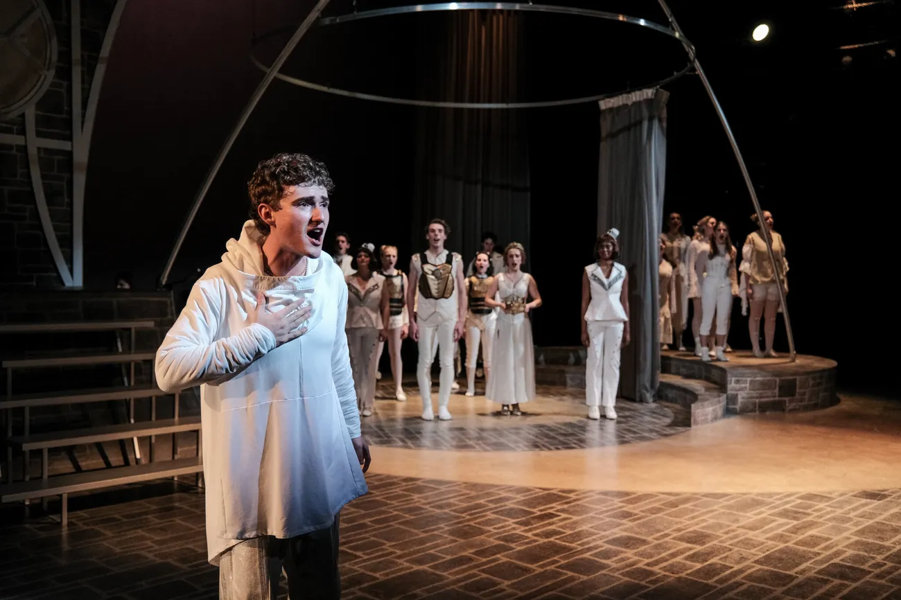 An actor sings on stage in the foreground. In the background is a cast of actors standing and listening. All are in costume.