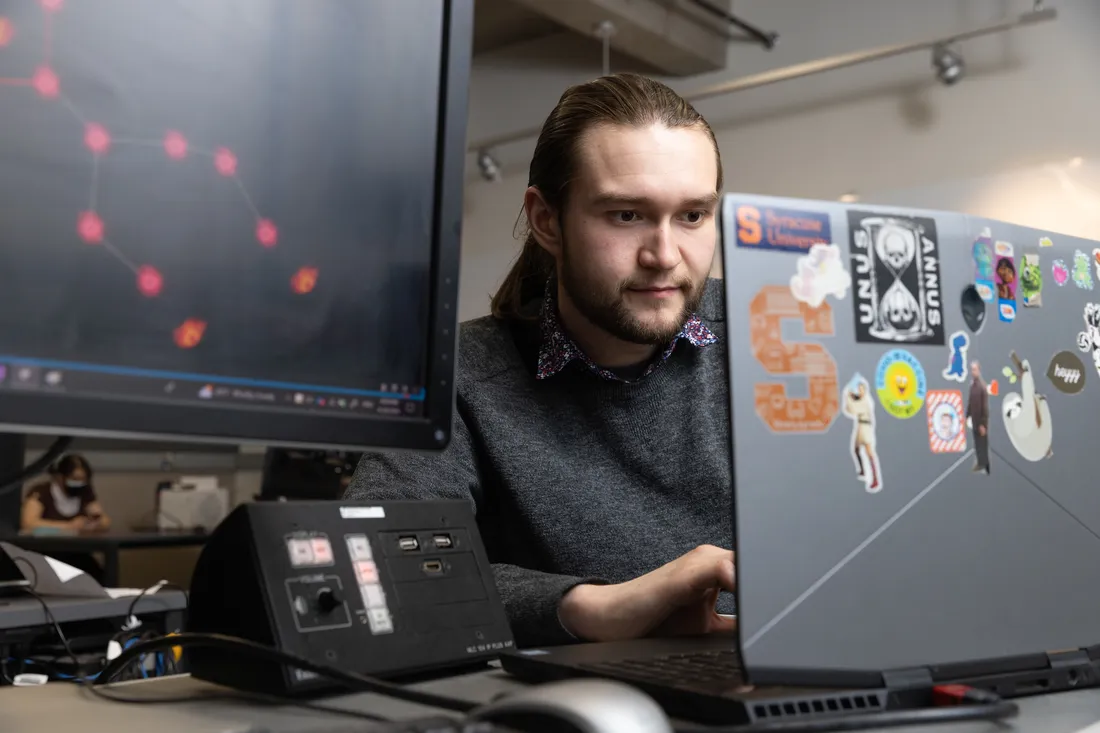 A student works on a laptop and sits next to a computer screen displaying a computer game.