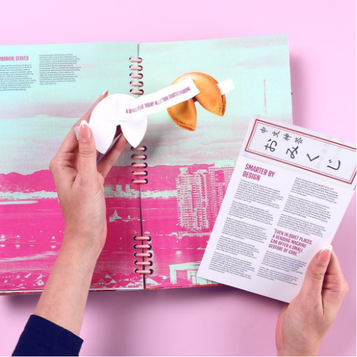 A person holds a paper fortune cookie message above an open, brightly colored spiral-bound magazine spread, while another hand holds a printed card with Chinese characters and design text. The scene is styled with pink and pastel tones.