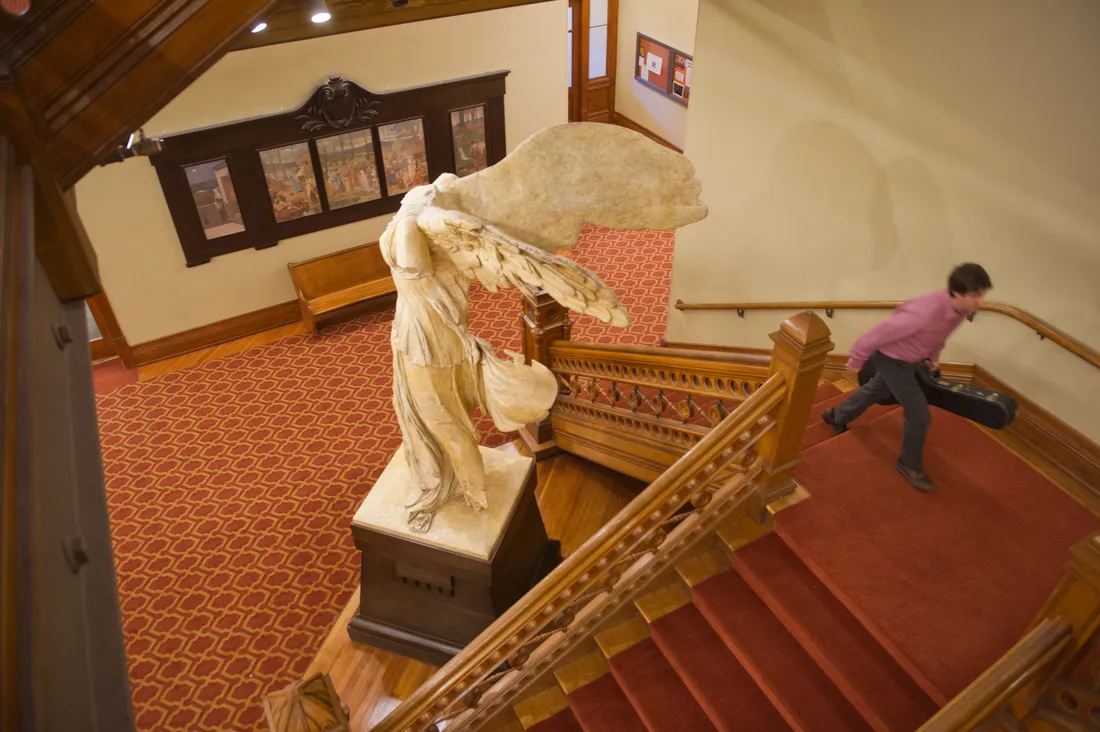 A person walks up a staircase in an old, historic building. Next to the staircase is a statue, "Winged Victory."