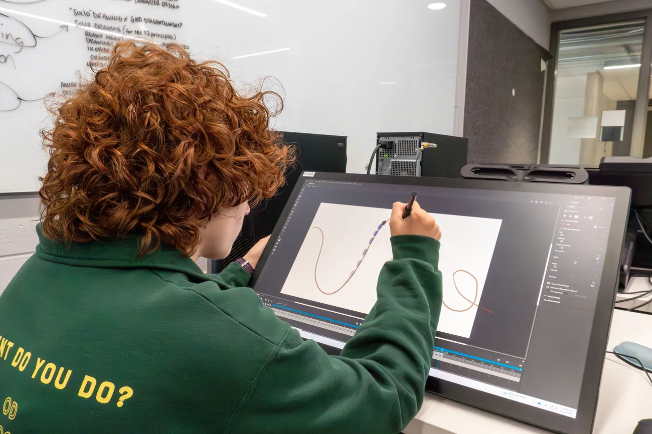 A student sits at a computer in a computer lab and uses a stylo to draw curved lines on the screen.
