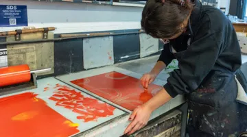 A person uses orange-red paint in the printmaking area.