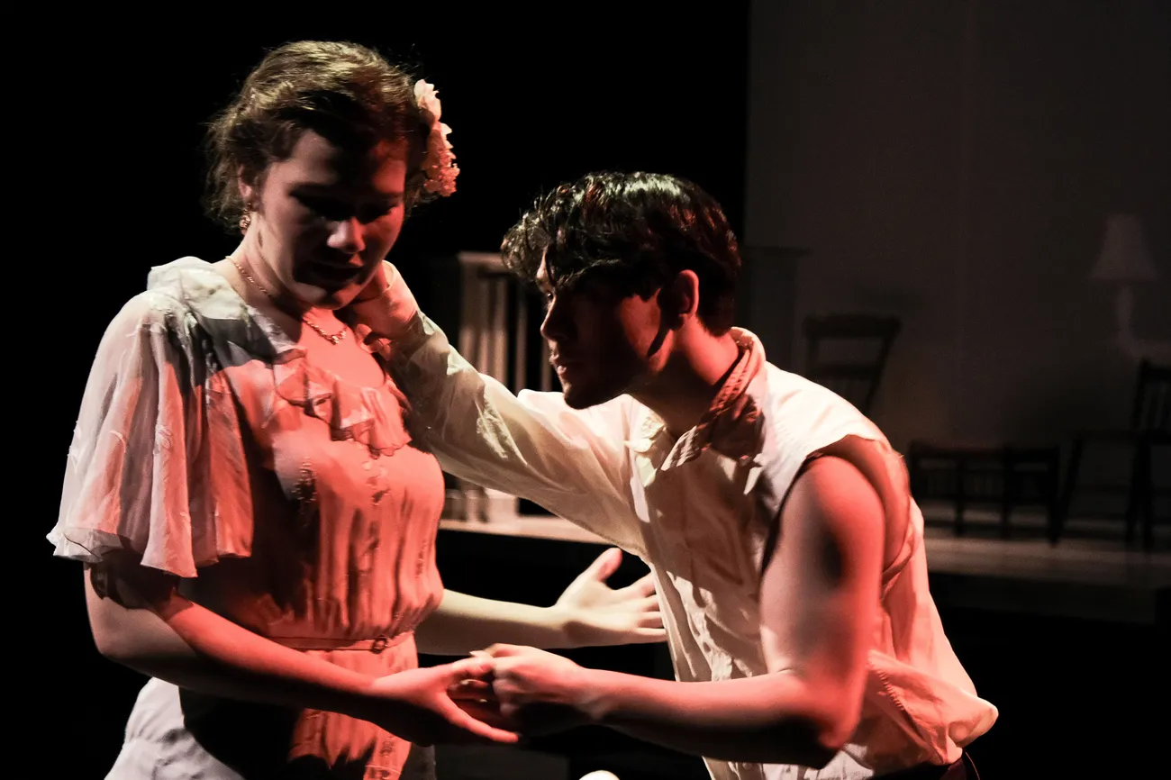 Two actors in costume act on a dimly lit stage. One actor grasps the other's hand and shoulder.