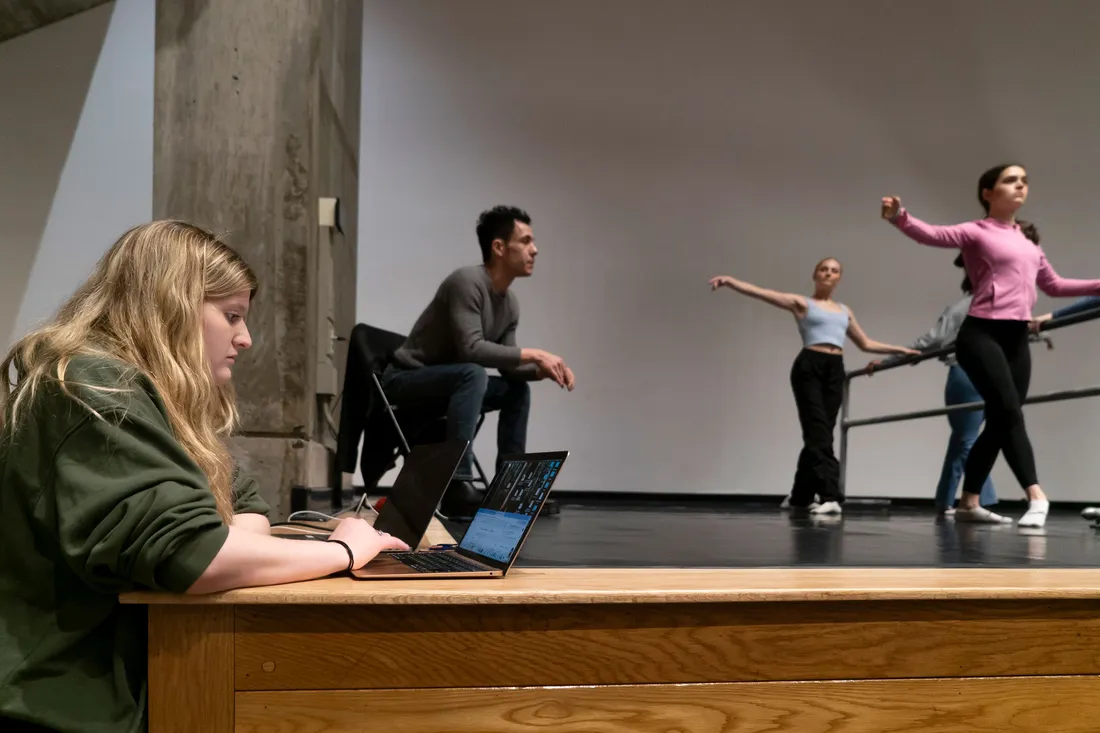 A student stands next to a stage using a laptop that rests on it. In the background is a director and dancers having a rehearsal.