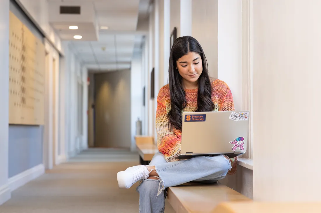 A student sits on a bench looking at a laptop.